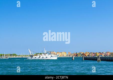 Chioggia, Italia, 16 settembre 2019: Nave da pesca in acque lagunari con fila di edifici colorati della città di Sottomarina, cielo blu di fondo nella giornata estiva, Regione Veneto Foto Stock