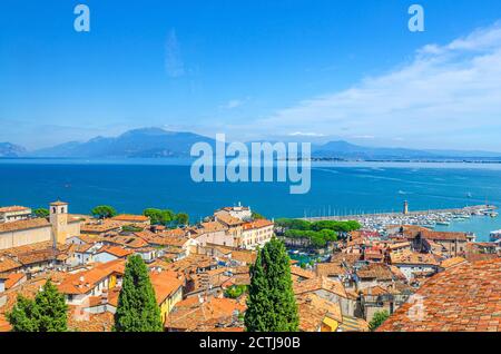 Vista panoramica aerea della città di Desenzano del Garda, tetti in tegole rosse, superficie acquatica del Lago di Garda, catena montuosa del Monte Baldo, penisola di Sirmione, faro molo, Lombardia, Italia settentrionale Foto Stock