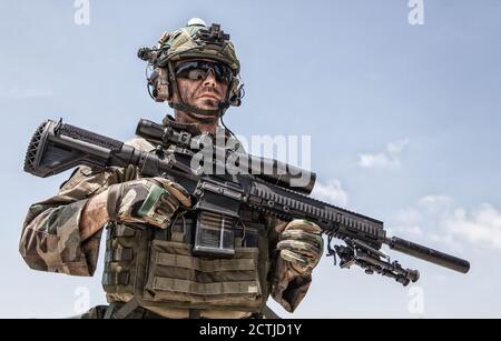 Ritratto a mezza lunghezza di cecchino o marcationista di forze speciali, soldato dell'esercito in uniforme camuffata e casco con cuffie radio, in piedi sullo sfondo del cielo blu, tenendo fucile da cecchino con campo ottico Foto Stock