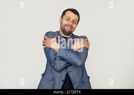 Giovane uomo d'affari con barba che si agguonava felice e positivo, sorridendo fiducioso. Auto-amore e cura di sé. Ritratto di un uomo su sfondo grigio. Foto Stock