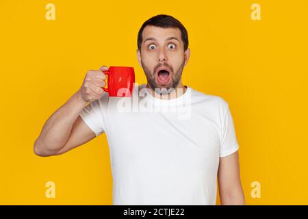 Il giovane con barba in una t-shirt bianca beve una tazza di caffè, spaventato con un volto sorpreso, spaventato ed eccitato da un'espressione di paura. Si trova su uno sfondo giallo isolato. Foto Stock