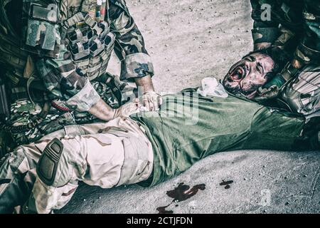 Soldati che cercano di smettere di sanguinare contro il compagno ferito che si trova a terra, sofferente e urlando a paine. Comando combattente premendo con le mani su ferita sanguinosa allo stomaco degli amici, dando assistenza di emergenza Foto Stock