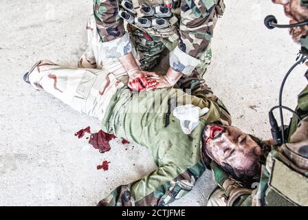 Soldati che cercano di smettere di sanguinare contro il compagno ferito che si trova a terra, sofferente e urlando a paine. Comando combattente premendo con le mani su ferita sanguinosa allo stomaco degli amici, dando assistenza di emergenza Foto Stock