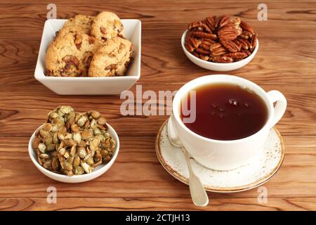 Biscotti d'avena con noci e uvetta, biscotti con semi di zucca ricoperti di zucchero, noci pecan in un recipiente e una tazza di tè nero su un tavolo di legno. Primo piano Foto Stock