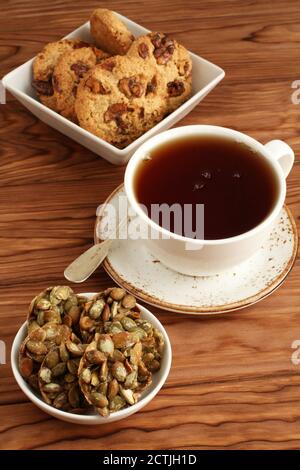 Biscotti d'avena con noci e uvetta, biscotti con semi di zucca ricoperti di zucchero in una ciotola e una tazza di tè nero su un tavolo di legno. Primo piano Foto Stock