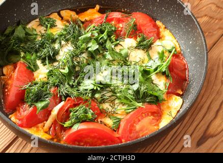 Uova strapazzate con pomodori, cosparse di aneto tritato e prezzemolo in una padella di teflon su un tavolo di legno marrone. Primo piano Foto Stock