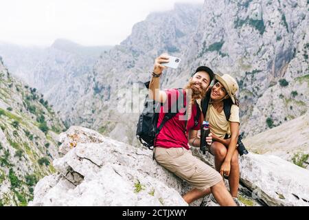 Coppia sorridente che prende selfie mentre si siede sulla roccia a Ruta del Cares, Asturias, Spagna Foto Stock