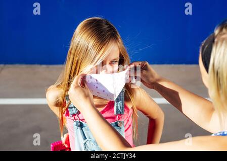 Madre che mette la maschera protettiva sulla faccia della figlia durante la giornata di sole Foto Stock