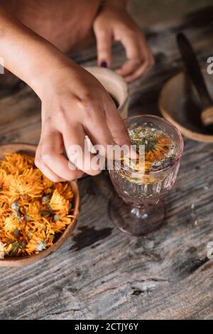 Mano di donna che prepara tè fresco alle erbe con fiore dentro vetro su tavolo di legno Foto Stock