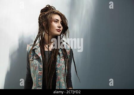 Giovane donna hipster con lunghi dreadlock in piedi contro parete grigia Foto Stock