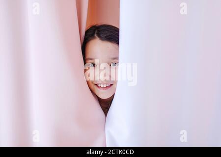 Ragazza sorridente che si nasconde dietro la tenda a casa Foto Stock