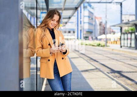 Donna sorridente che usa lo smartphone mentre aspetta alla stazione del tram Foto Stock