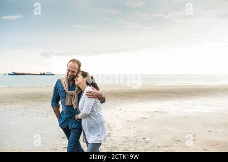 Uomo maturo abbracciando la donna mentre camminando in spiaggia contro nuvoloso cielo Foto Stock