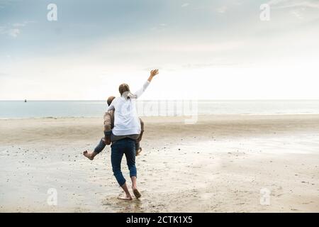 Uomo maturo piggybacking donna allegra mentre camminando in spiaggia durante tramonto Foto Stock