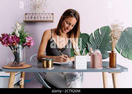 Donna sorridente che scrive nel diario mentre si siede al tavolo contro parete Foto Stock