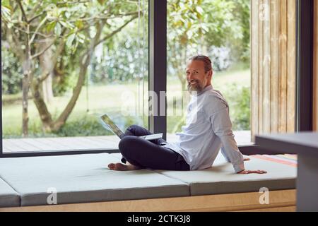 Sorridente uomo maturo con il portatore che usa il computer portatile mentre si siede a letto dalla finestra a casa Foto Stock