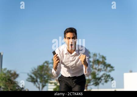 Uomo d'affari eccitato che tiene lo smartphone urlando mentre si levano in piedi contro chiaro cielo blu Foto Stock