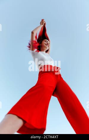 Giovane donna sorridente che tira i capelli rossi tinti in piedi contro il cielo limpido Foto Stock