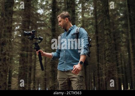Uomo che riprende con la macchina fotografica e il gimbal mentre si levano in piedi contro gli alberi in foresta Foto Stock