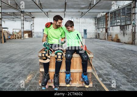 Felice ragazzo con braccio intorno al padre mentre si siede con hockey bastoni su scatola di legno a corte Foto Stock