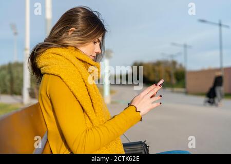 Giovane donna che si sta testando attraverso il telefono cellulare mentre si siede sulla panca nel parco Foto Stock