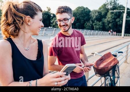Felice coppia che usa gli smartphone mentre si sta in piedi sul ponte a. tramonto Foto Stock