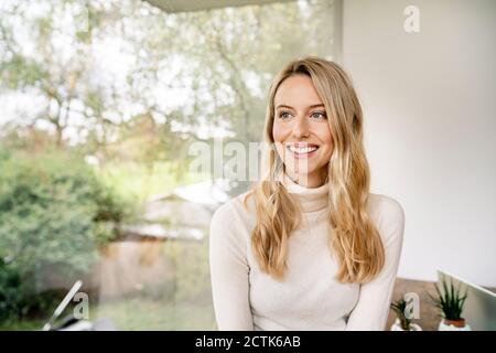 Sorridente donna d'affari premurosa con i capelli biondi seduti dalla finestra dentro ufficio Foto Stock