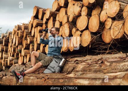 Escursionista maschile guardando attraverso binocolo mentre si siede sul log contro cumulo di legno in foresta Foto Stock