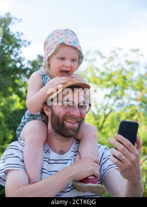 Figlia che si stacca la lingua mentre il padre prende selfie su intelligente telefono Foto Stock