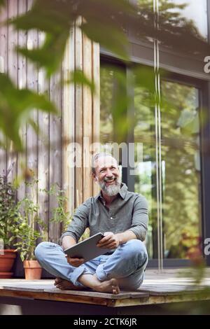 Uomo sorridente con un tablet digitale che guarda lontano mentre si è seduti fuori casa piccola Foto Stock