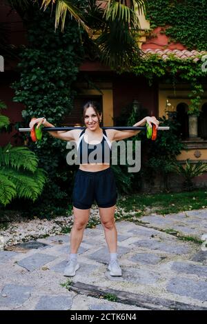 Sorridente donna adulta media che solleva pesi mentre si è in piedi in cortile Foto Stock