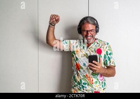 Uomo sorridente con la mano sollevata utilizzando il telefono cellulare contro armadio Foto Stock