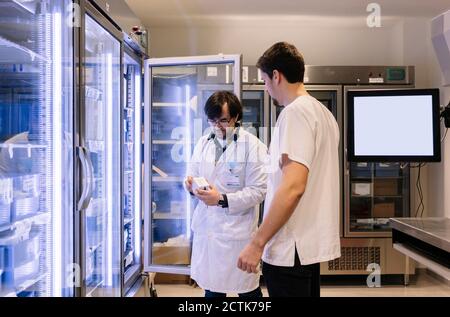 I farmacisti maschili che prendono l'inventario mentre si levano in piedi dal frigorifero illuminato dentro ospedale Foto Stock