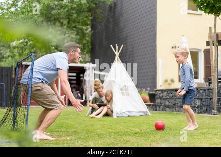 Uomo e figlio giocano a calcio mentre la donna si siede con la figlia in tenda al cortile posteriore Foto Stock