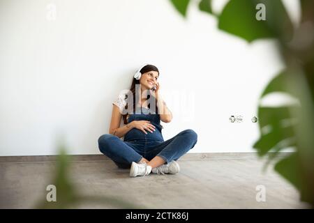 Donna incinta sorridente che ascolta la musica attraverso le cuffie mentre si siede muro in nuova casa Foto Stock