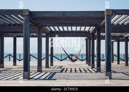 Donna sdraiata in amaca appesa a struttura metallica sul lungomare durante il giorno di sole Foto Stock