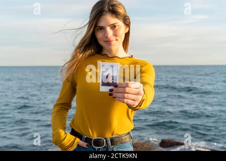 Donna sorridente che mostra una fotografia mentre si è in piedi contro il mare Foto Stock