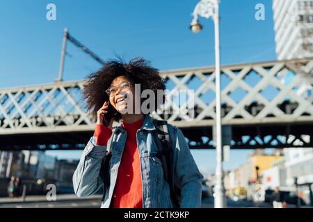 Giovane donna afro allegra che parla su smartphone mentre si è in piedi contro il ponte in città Foto Stock