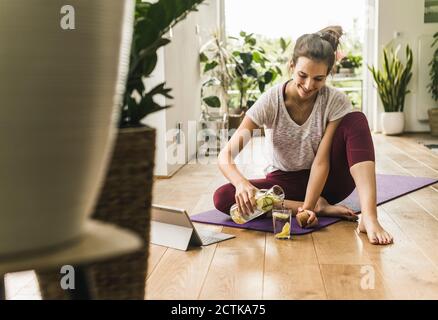 Sorridente giovane donna che versa in bicchiere mentre si siede tappetino per esercizi a casa Foto Stock