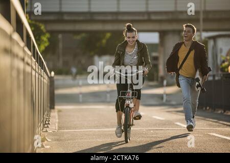 Ragazzo allegro che corre dietro girlfriend in bicicletta in città giorno di sole Foto Stock