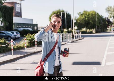 Un giovane sorridente che parla sul telefono cellulare mentre attraversa la strada in città Foto Stock