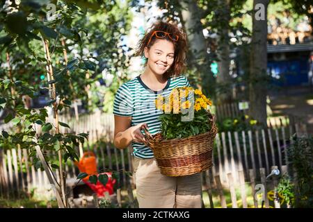 Giovane donna sorridente che tiene il cesto con fiori gialli mentre si è in piedi nel giardino posteriore del cortile Foto Stock