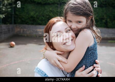 Ragazza abbracciando madre sorridente al campo di pallacanestro nel cortile posteriore Foto Stock