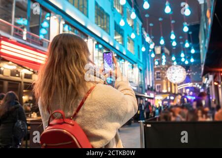 Giovane donna che fotografa luci di Natale illuminate appese in città a. notte Foto Stock