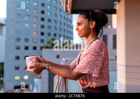 Bella donna che tiene la tazza di caffè mentre si sta in piedi in balcone godendo tramonto Foto Stock