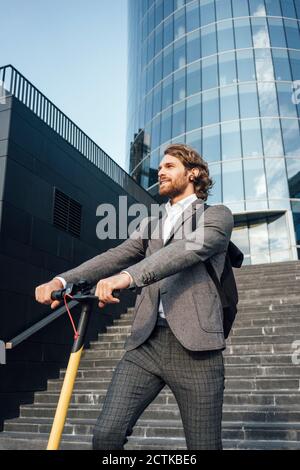 Bel professionista maschile che guarda via mentre si è in piedi con la spinta elettrica scooter contro l'edificio dell'ufficio Foto Stock