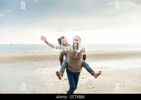 Felice uomo maturo piggybacking donna mentre camminando in spiaggia contro cielo nuvoloso Foto Stock