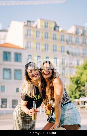 Allegre amiche che tengono bottiglie di birra mentre si levano in piedi contro l'edificio in città Foto Stock