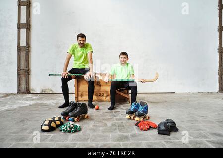 Padre e figlio seduti con bastoni da hockey su scatola di legno da attrezzature sportive contro il muro in campo Foto Stock