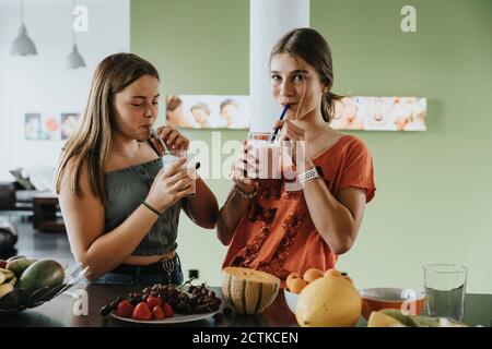 Ragazze adolescenti in piedi in cucina bevendo frullati di frutta fresca con cannucce da bere